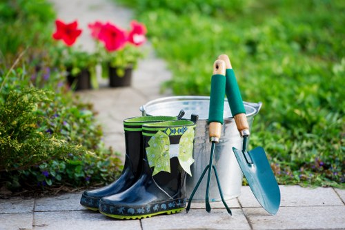 A pair of dark rubber gardening boots with green and white patterned socks draped inside, positioned on a paved pathway in a garden setting. Next to the boots, there is a white plastic bucket, a small hand trowel with a teal handle, and a long-handled gardening fork. In the background, vibrant pink flowering plants and lush green foliage can be seen, with a blurred garden path leading through well-maintained flower beds. The scene appears to be during daytime with natural lighting, suggesting a tidy outdoor space suitable for gardening activities, which aligns with services offered by Garden Maintenance Mill Hill in the local area. The overall environment reflects a well-kept garden yard with a mix of flowering plants, soil, and paved surfaces, emphasizing outdoor maintenance and gardening work in a typical residential garden in Mill Hill or nearby areas within the NW7 postcode.