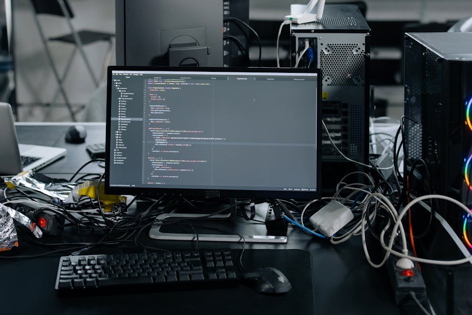 A cluttered workspace featuring a computer monitor displaying lines of code, surrounded by tangled cables, a keyboard, mouse, and assorted electronic components, with a laptop partially visible on the