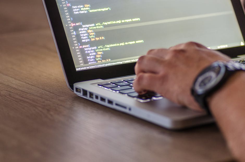 A close-up image shows a person's hand typing on a silver laptop with a black keyboard, placed on a wooden surface. The laptop screen displays lines of colorful code, indicating a programming or websi