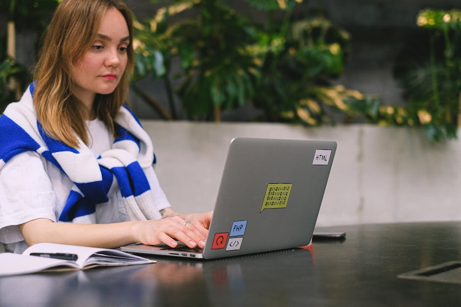 A young woman with shoulder-length light brown hair is sitting outdoors at a black table in a garden setting, working on a silver laptop decorated with colorful stickers labeled 'HTML,' 'PHP,' and one